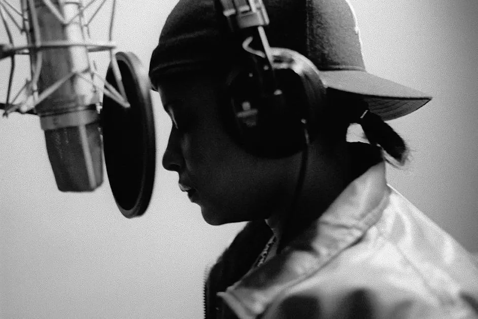 Black-and-white side profile of a young person wearing a backwards cap and large headphones, standing in front of a studio microphone with a pop filter.